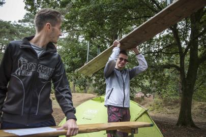 Twee jongemannen in een bos, één houdt een plank vast en de ander tilt een plank boven zijn hoofd. Een groene tent staat op de achtergrond.