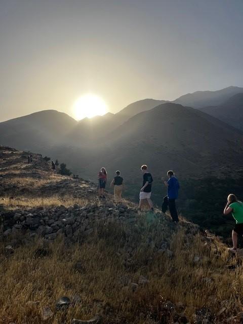 Vijf jongeren aan het wandelen op een berg, met meer bergen en de zonsondergang op de achtergrond