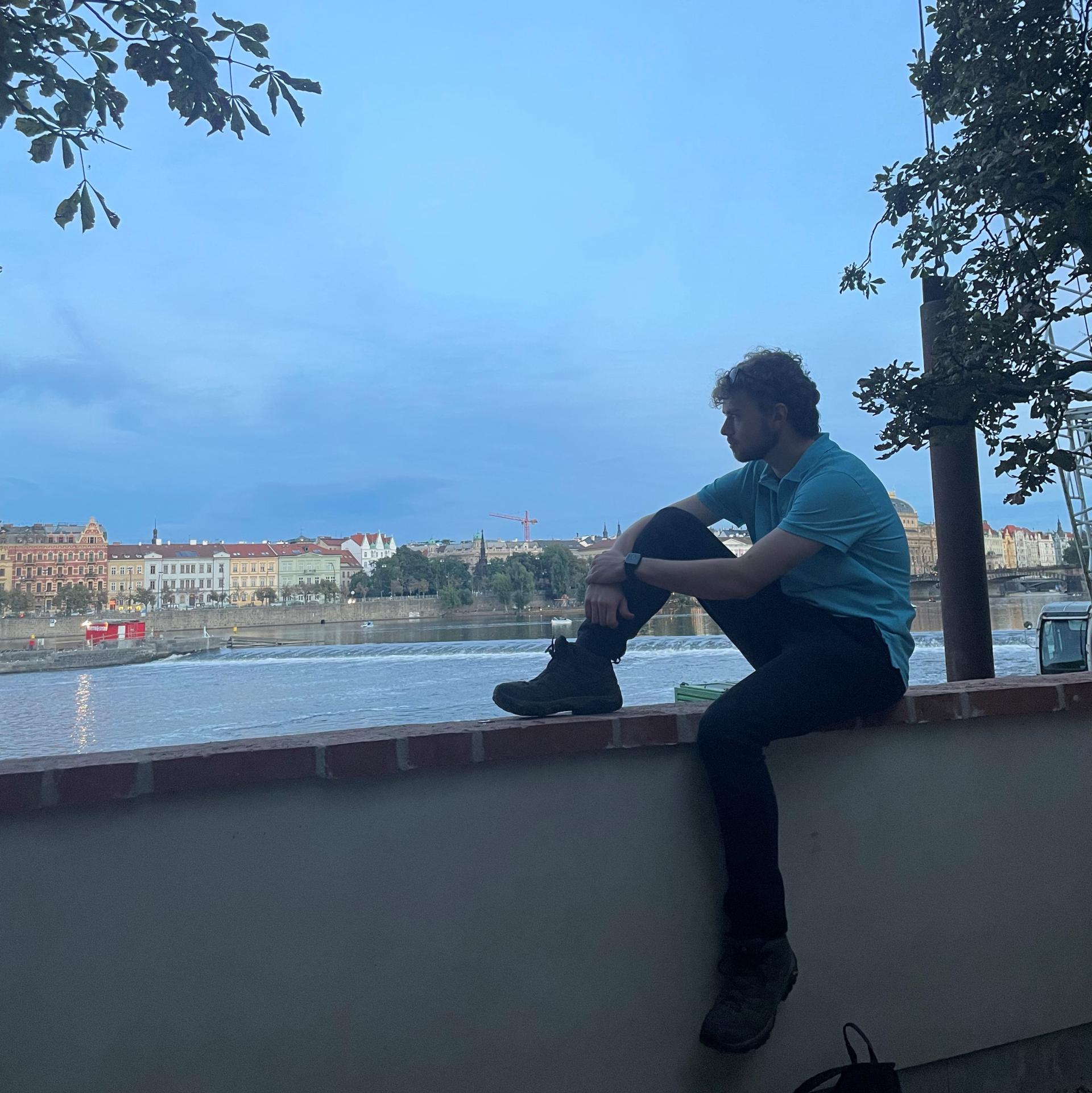 Luís zit met één been opgetrokken op een muurtje. Hij kijkt naar het uitzicht op de stad, met daarvoor een rivier.