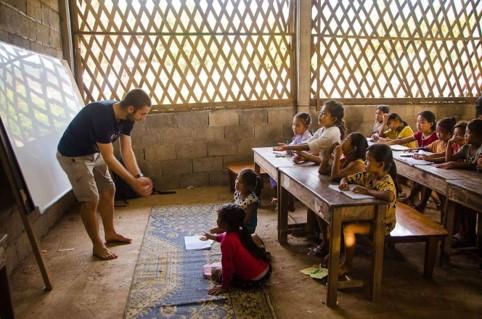 Een klas waar het licht binnenvalt door de ramen. Timo staat links aan het schoolbord en geeft les een de kindjes die rechts op de foto op houten schoolbanken zitten
