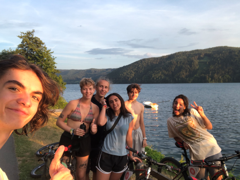 Mauro (links in beeld) neemt een selfie met vier andere jongeren voor een meer omringt met bergen. Ze poseren lachend voor de foto. De jongeren hebben fietsen bij. De zon schijnt.
