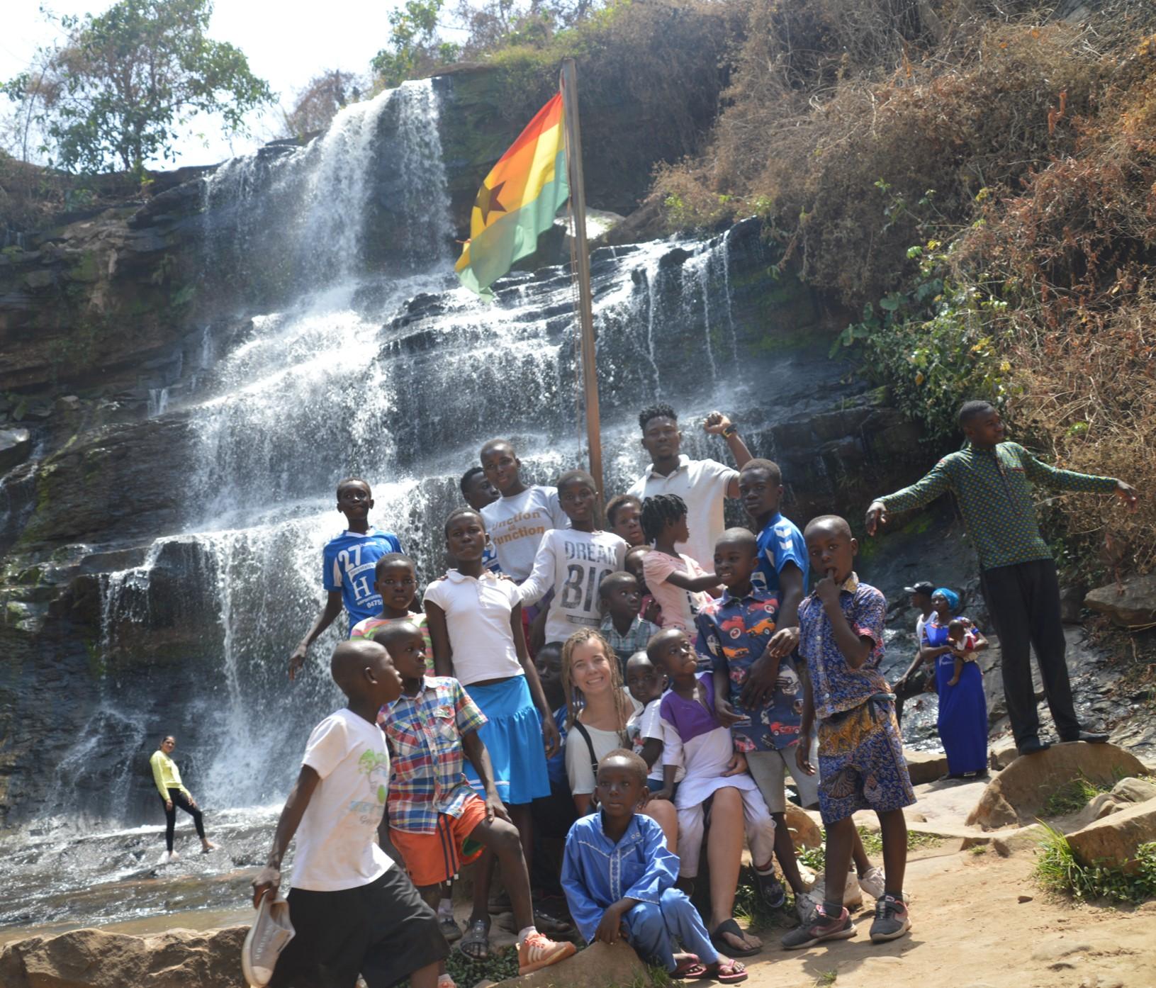 Melissa met een groep van ongeveer vijftien kinderen poserend met een waterval op de achtergrond. Vlak acht hen staat een vlaggenmast waaraan de Ghanese vlag wappert.