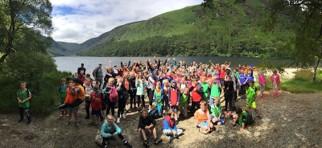 een grote groep kinderen poseert voor de foto. Ze staan in de natuur met een meer en bergen op de achtergrond.