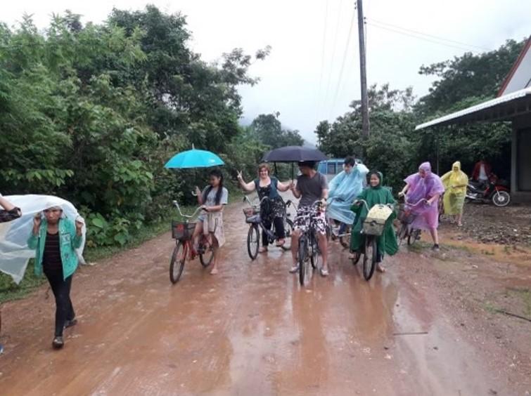 Een vijftal mensen poserend op de fiets in de regen op een modderige weg. Ze dragen regenkledij en paraplu's