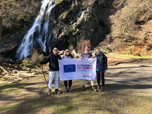 Willem poseert met drie andere jongeren voor een foto. Samen houden ze een vlag voor hen waarop het logo van het European Solidarity Corps staat. Ze staan in de natuur, met op de achtergrond een klein waterval.