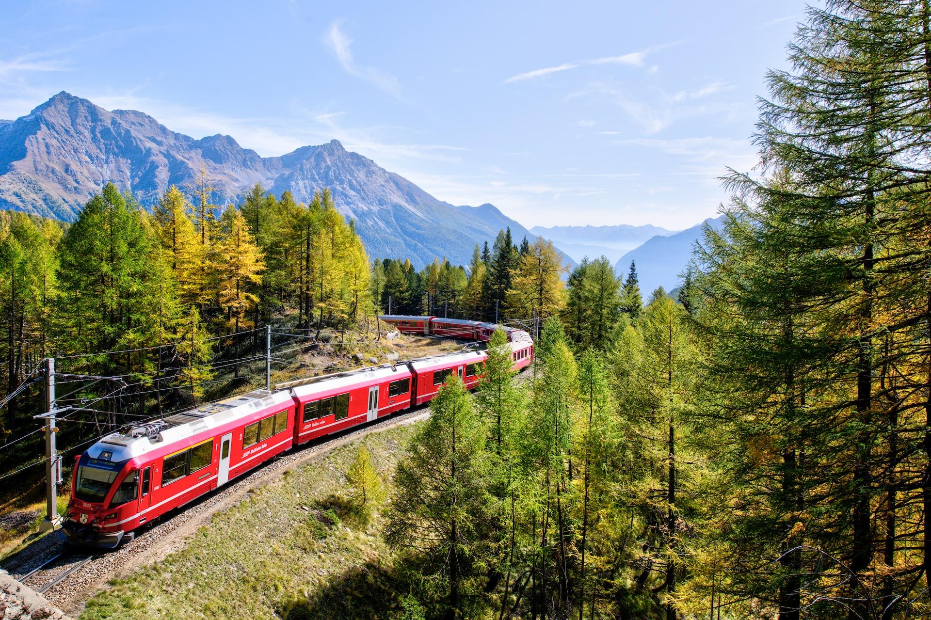Rode trein rijdt over het spoor tussen de naaldbomen. Het einde van de trein is niet zichtbaar in de bocht. Op de achtergrond zijn hoge bergen te zien. De zon schijnt.