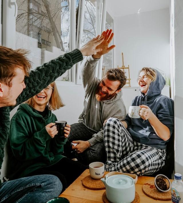 Vier mensen drinken koffie in een huiselijke setting. Ze lachen, twee van hen geven een high-five.